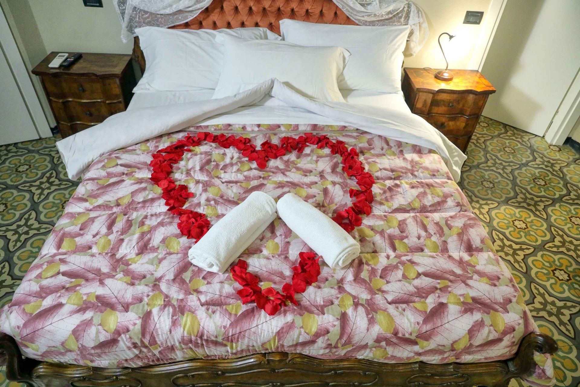 Bed with heart-shaped arrangement of red rose petals and rolled towels on floral bedspread.