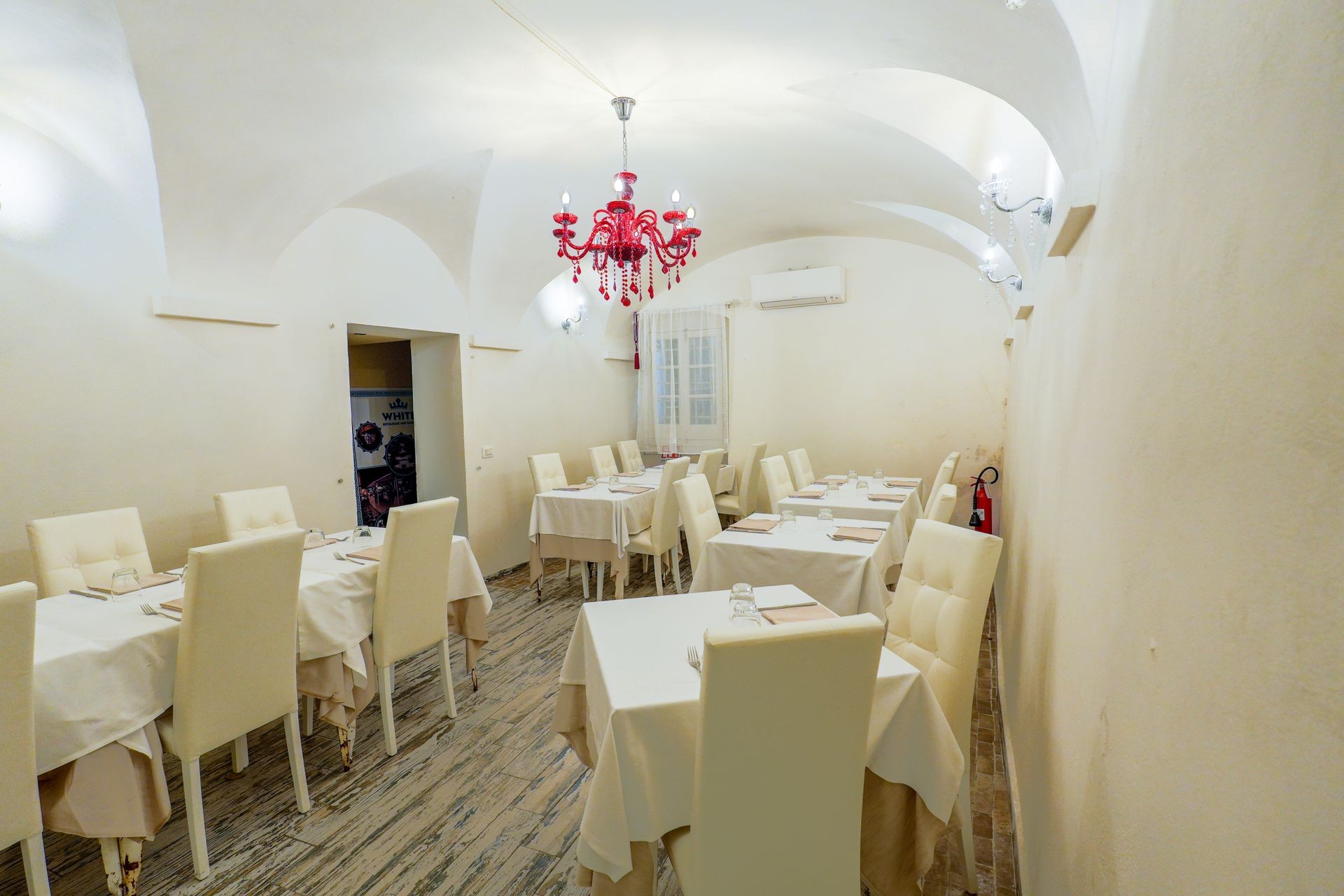 Elegant dining room with white tables and chairs, red chandelier, and arched ceiling.